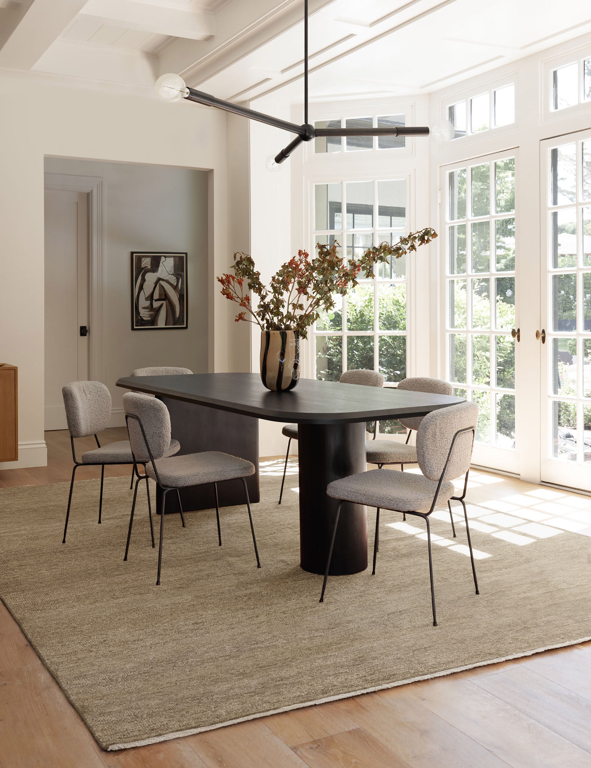 The Heritage wheat rug lays in a dining room with wrap-around windows, a black dining table, and upholstered dining chairs