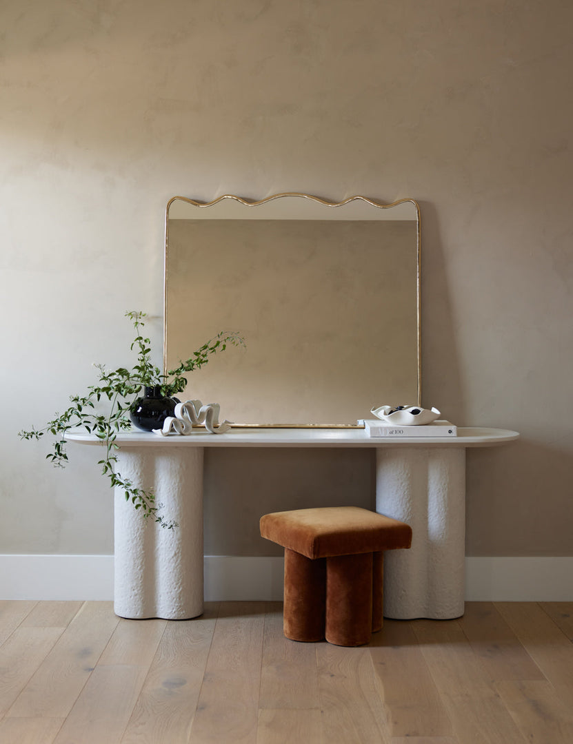 #showall::41056638140515 | The rook mantel mirror sits atop a white sideboard with pedestal legs next to a cognac velvet ottoman