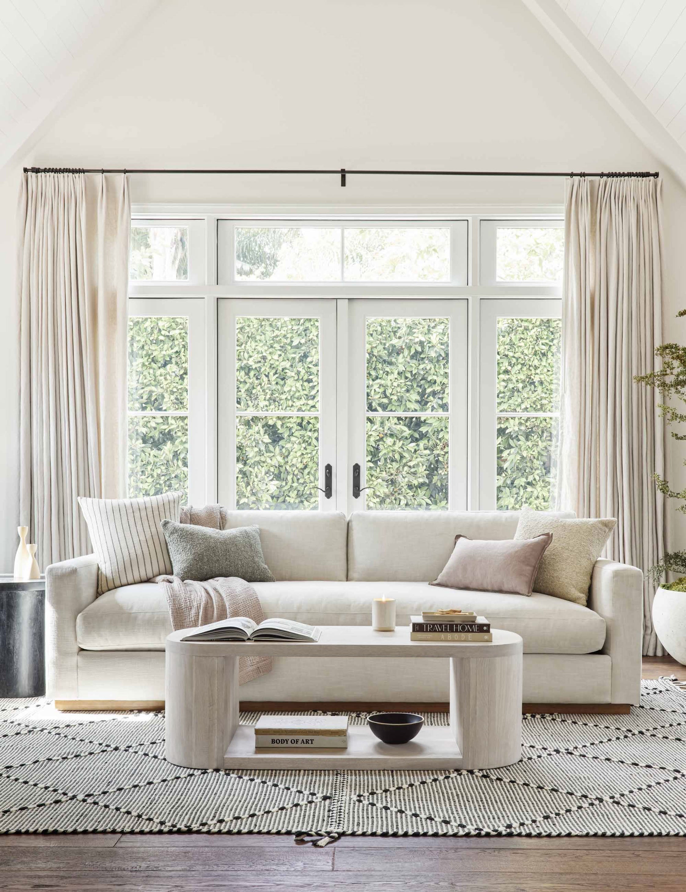 The Luna white-washed oak oval coffee table sits in a well-lit living room atop a white and black patterned rug in front of a white couch with long windows and french doors in the background.