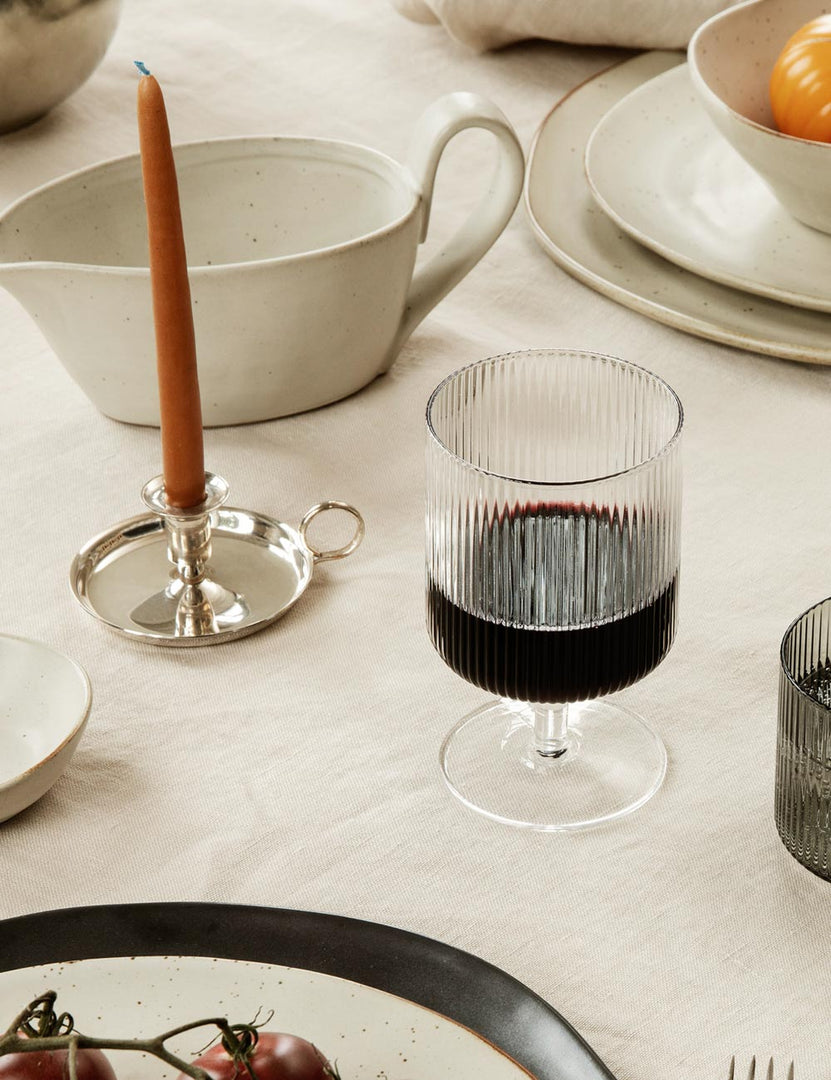 #color::clear #style::wine-glasses--set-of-2 | One rian ripple wine glass sits atop a dining room table with a terracotta taper candle and a white gravy boat
