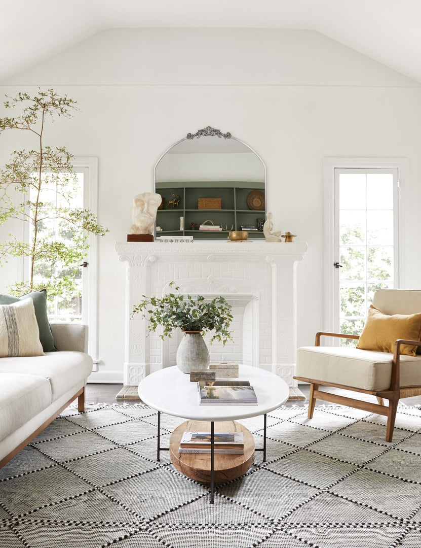 #color::silver | The Tulca arched silver mirror with flat bottom edge and traditional scroll detailing sits atop a fireplace in a bright living room with a natural sofa and accent chair and a black and white patterned rug.