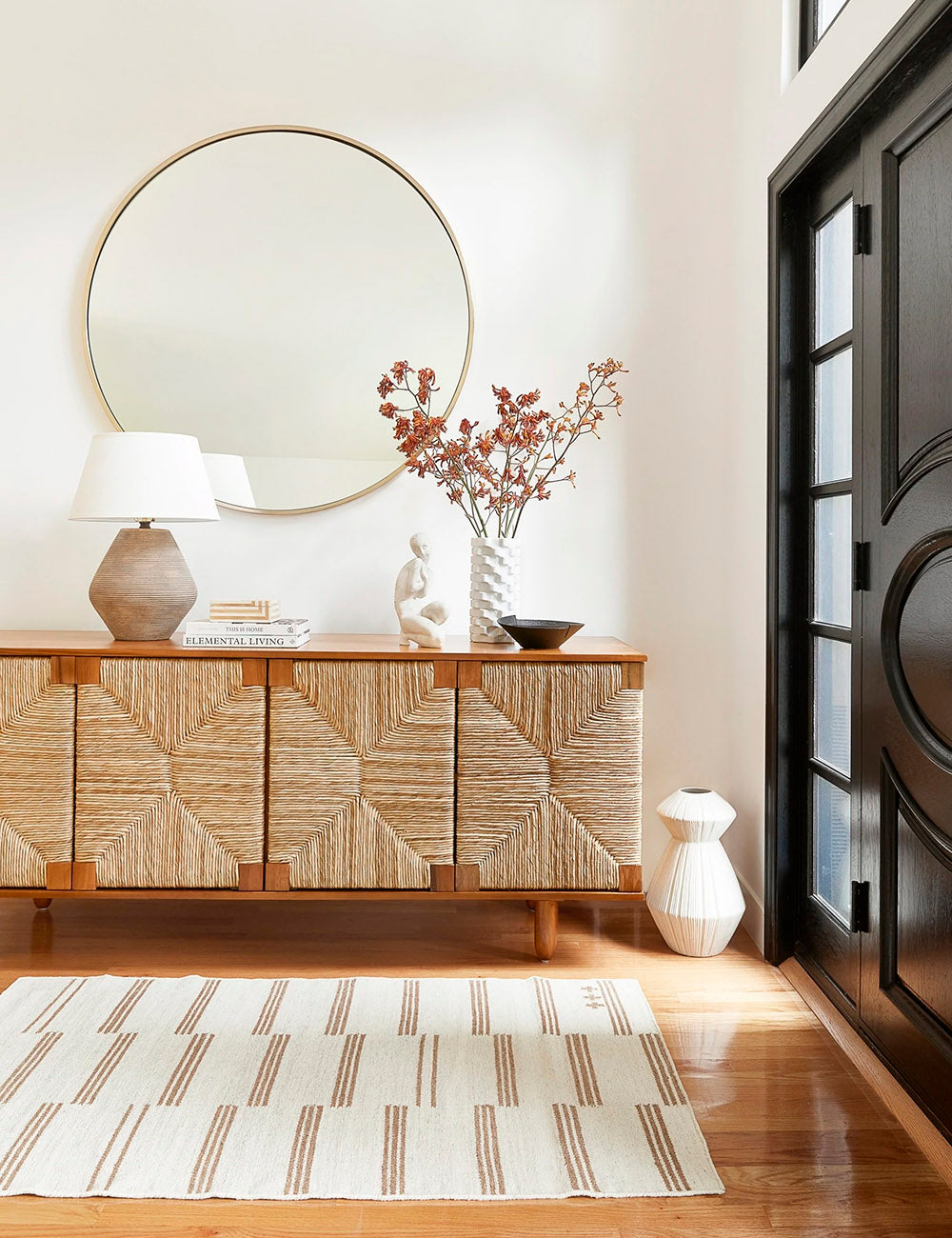 The Stripe break flatweave rug by Sarah Sherman Samuel lays in an entryway with a golden framed circular mirror, a woven and wooden sideboard, and various sculptural decorative objects