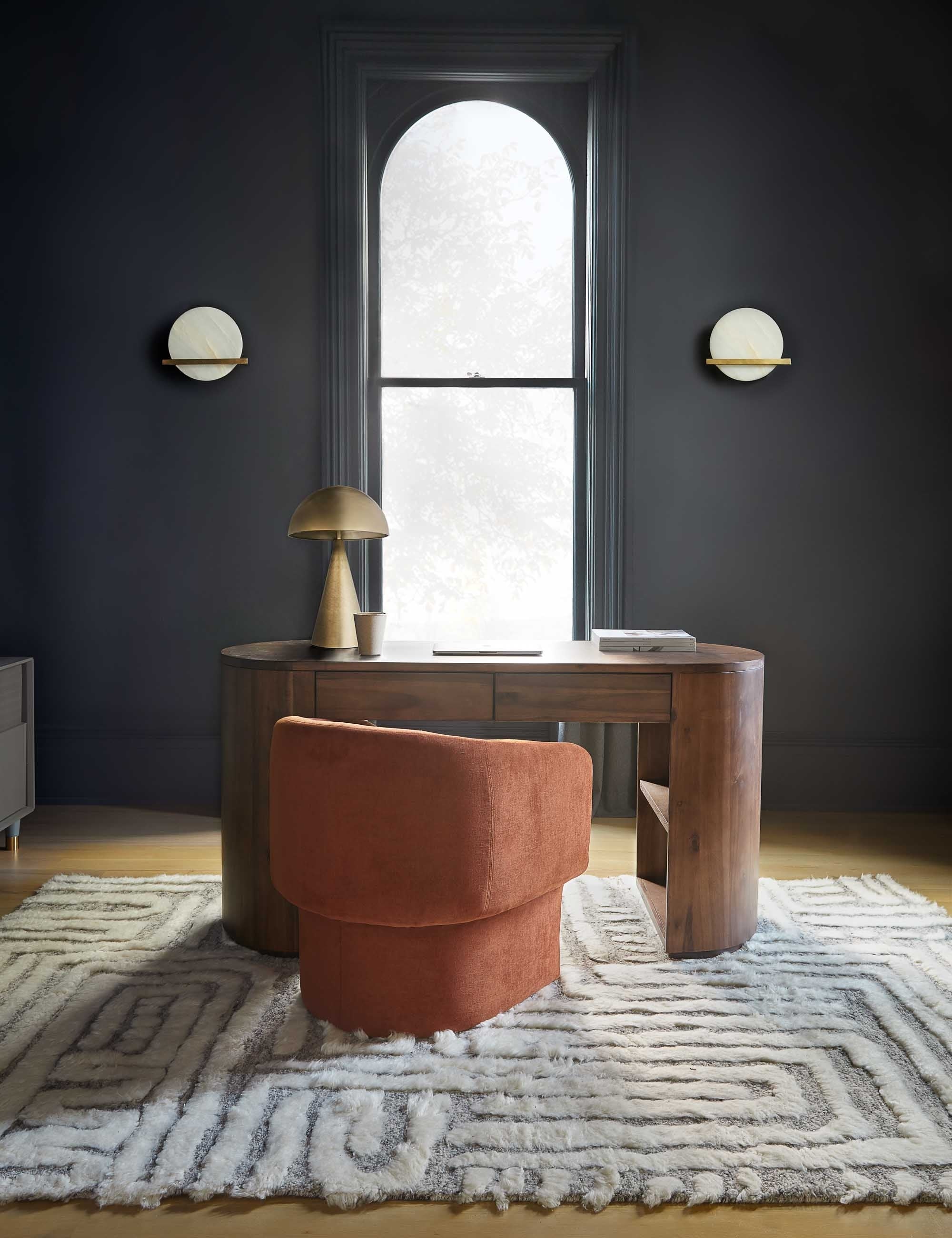 The braeburn rug lays in an office with black walls and a narrow central window under a desk and a rust orange chair