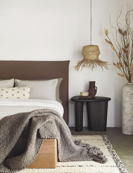 #color::mink | The Olema mink gray handwoven throw with fringed ends lays in a bedroom with a cream and black plush rug, a wooden-base bed, and a black sculptural nightstand 