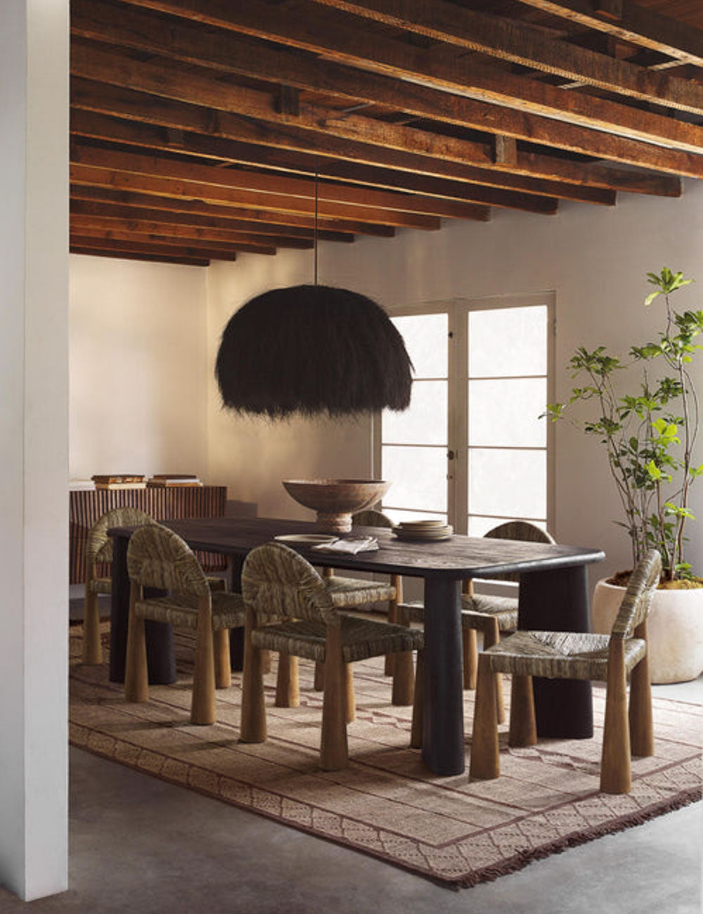 The Paige black plush pendant light with palm tree fibers sits in a dining room with wooden beamed walls, jute dining chairs, and a brown patterned rug