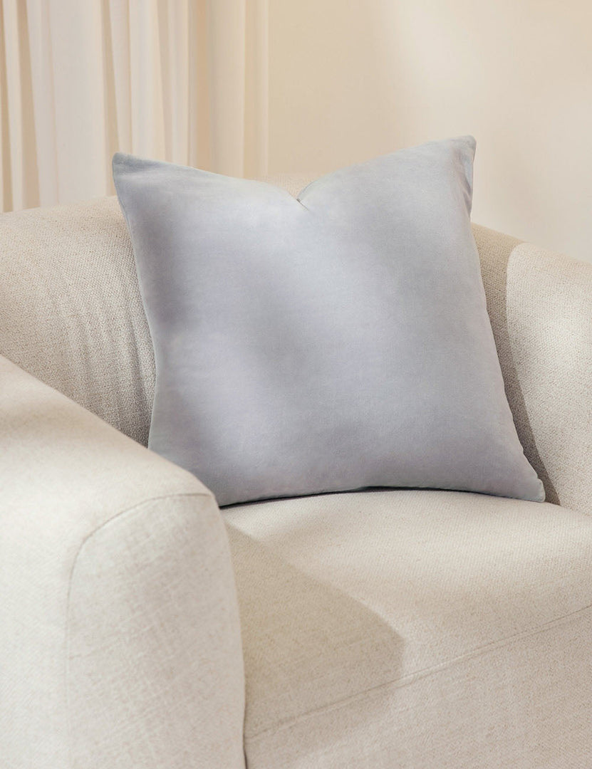 #color::ice-blue #size::20--x-20- #style::square  #fill type::with-feather-down-insert  #fill type::pillow-cover-only |   Charlotte ice blue square velvet pillow sits on a white accent chair with a white curtain in the background