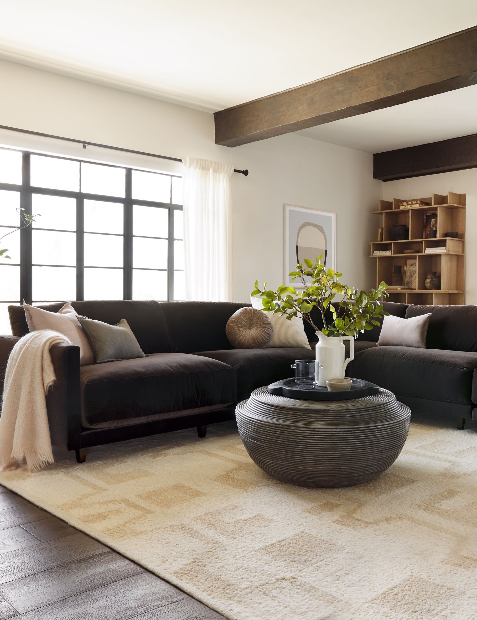 The Monroe warm gray velvet round pillow sits on a brown sectional in a living room with a round coffee table and a patterned rug