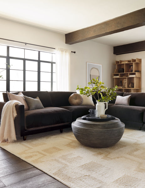 #color::warm-gray | The Monroe warm gray velvet round pillow sits on a brown sectional in a living room with a round coffee table and a patterned rug