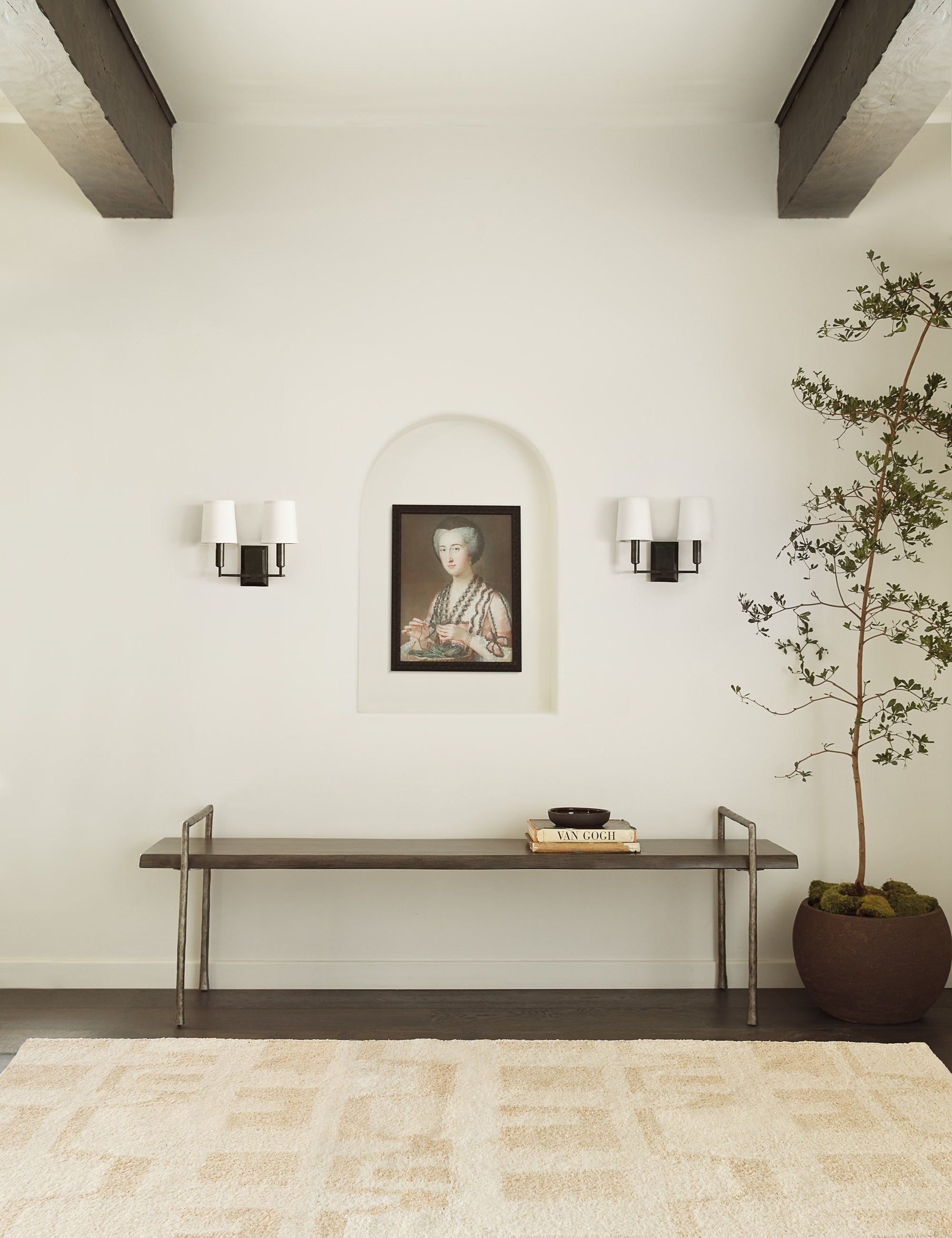 The Charlie dark bronze double sconce with parchment shades and angular arms is mounted in an entry way with a natural-toned plush rug, a portrait wall art, and long metal bench