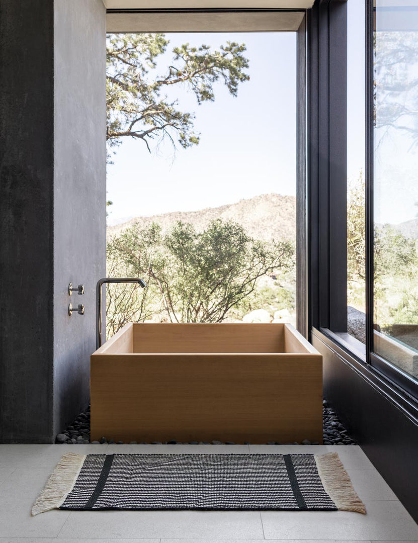 #size::1-9--x-2-10- #size::2--x-5- | The Ines handwoven black-and-white striped mat lays on a gray tiled floor in a bathroom with a wooden square bathtub and floor to ceiling windows