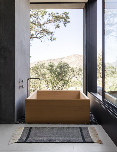 #size::1-9--x-2-10- #size::2--x-5- | The Ines handwoven black-and-white striped mat lays on a gray tiled floor in a bathroom with a wooden square bathtub and floor to ceiling windows