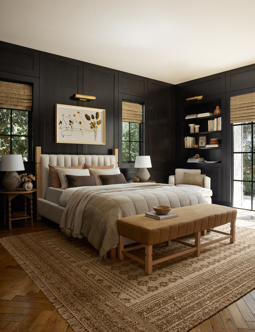 #showall::40199323779171 | The Ambleside camel linen bench sits atop a patterned rug in a room with accented black wooden walls and a natural linen bed