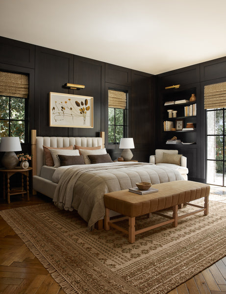 #showall::40199323779171 | The Ambleside camel linen bench sits atop a patterned rug in a room with accented black wooden walls and a natural linen bed