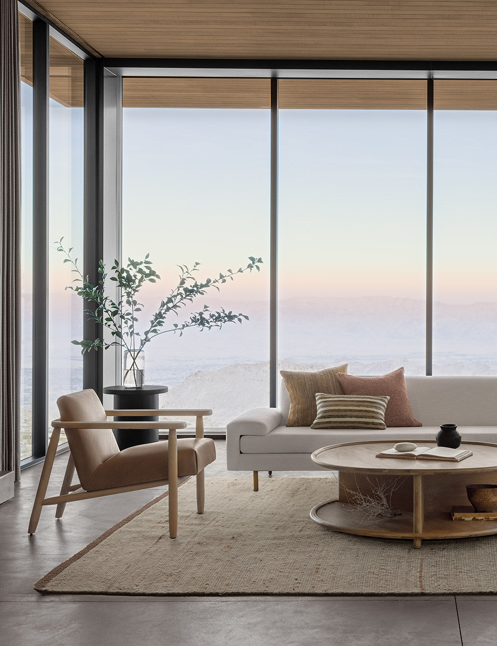 The Khloe natural rug lays in a living room with wrap around floor to ceiling windows under an ivory sofa