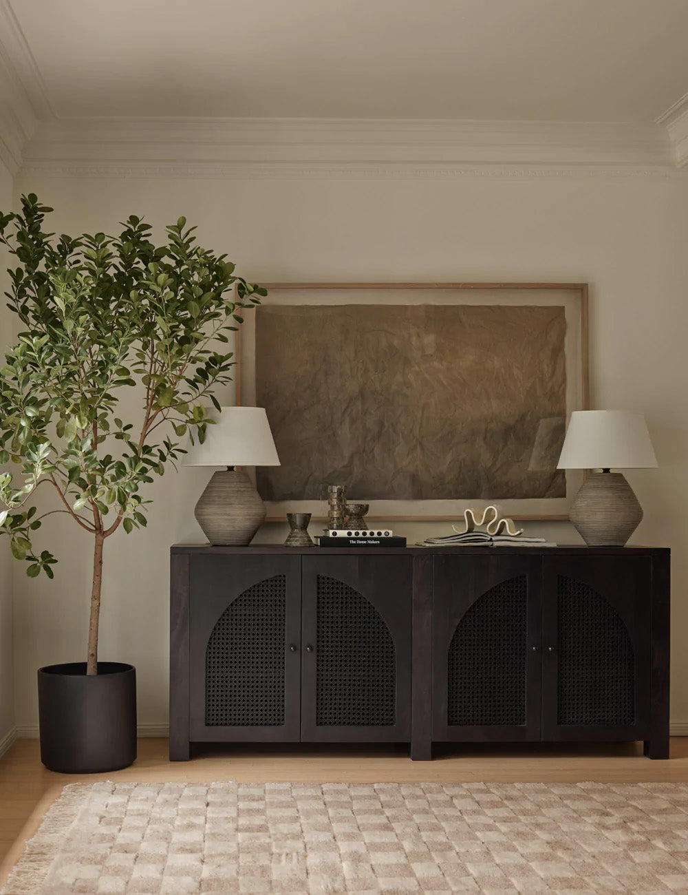The Irregular beige checkerboard rug by Sarah Sherman Samuel sits in a room with a black wooden sideboard, a large brown wall art, and sculptural stone lamps.