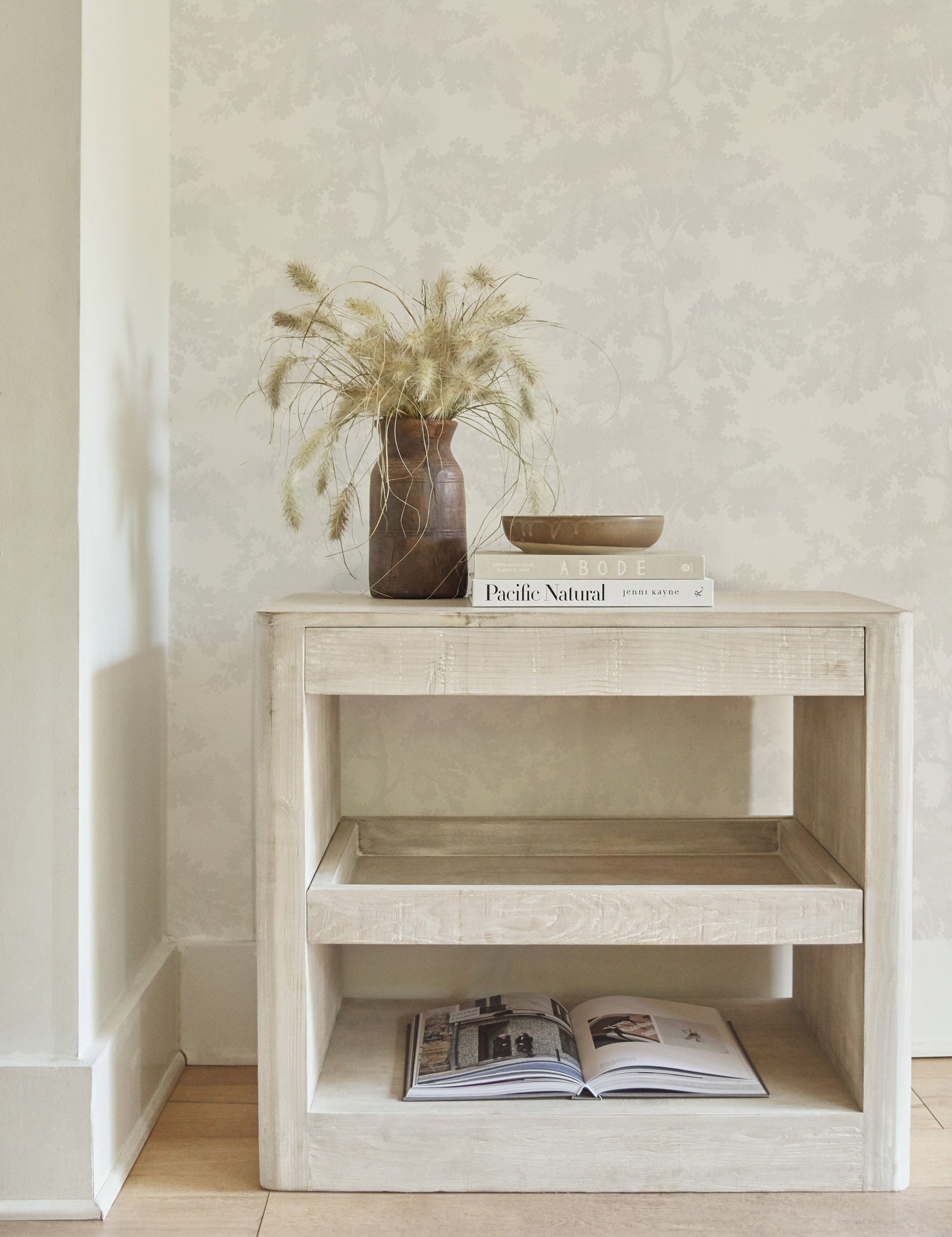 The Scalamandre botanical inspired white raphael wallpaper is in a room with a whitewashed side table with books sitting atop it