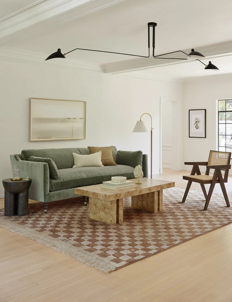 #color::ochre #size::2-6--x-8- #size::6--x-9- #size::8--x-10- #size::9--x-12- #size::10--x-14- #size::12--x-15- | The Irregular ochre checkerboard rug by Sarah Sherman Samuel sits in a living room with a sage velvet couch, a burl wood coffee table, and a woven cane accent chair.