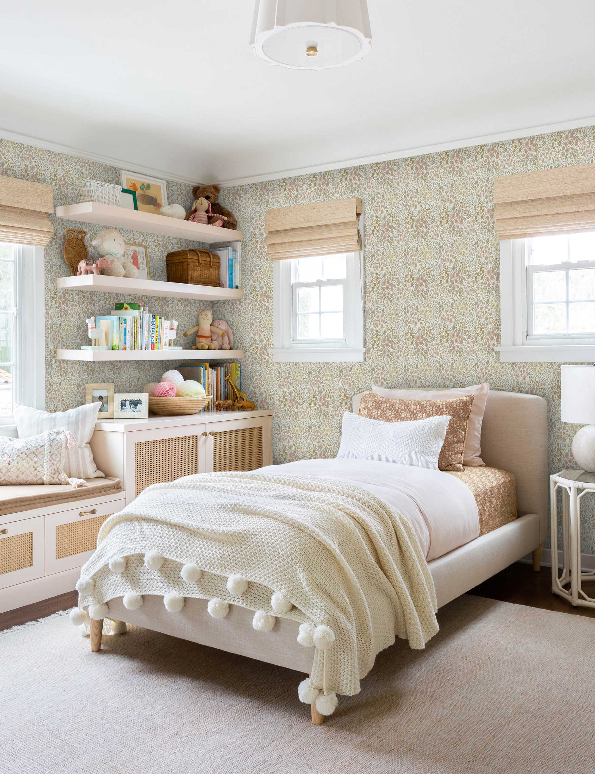 Warm-toned Floral Field Wallpaper by Rylee + Cru is in a child's bedroom with a toy-filled shelf and a bed with a pom pom throw blanket
