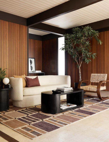 #color::black | The Luna black wood oval coffee table sits in a retro living room atop a burgundy and orange patterned rug surrounded by a cream boucle rounded couch and a woven accent chair with wooden arms and legs.