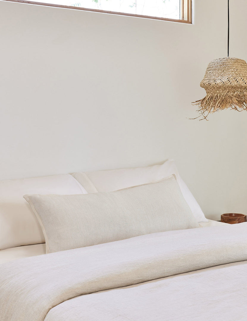 #color::ivory #style::extra-long #fill type::with-feather-down-insert #fill type::pillow-cover-only | The arlo Ivory long lumbar pillow lays on a bed with ivory linens with a jute pendant light hanging next to it