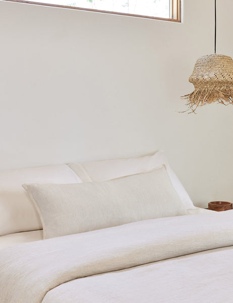 #color::ivory #style::extra-long #fill type::with-feather-down-insert #fill type::pillow-cover-only | The arlo Ivory long lumbar pillow lays on a bed with ivory linens with a jute pendant light hanging next to it