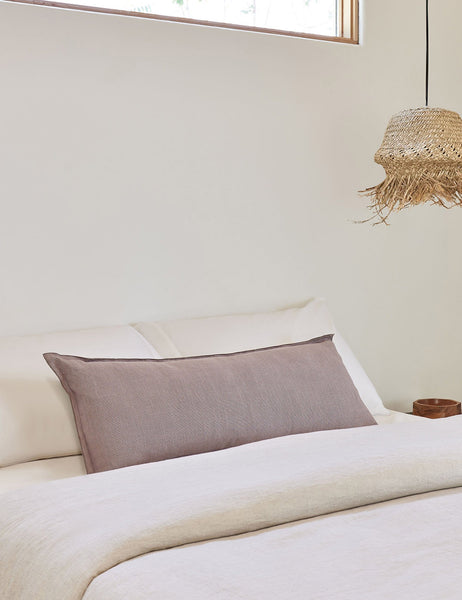 #color::dark-natural #style::extra-long #fill type::with-feather-down-insert #fill type::pillow-cover-only | The arlo Dark Natural long lumbar pillow lays on a bed with ivory linens with a jute pendant light hanging next to it