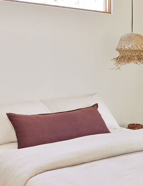 #color::aubergine #style::extra-long #fill type::with-feather-down-insert #fill type::pillow-cover-only | The arlo Aubergine burgundy long lumbar pillow lays on a bed with ivory linens with a jute pendant light hanging next to it