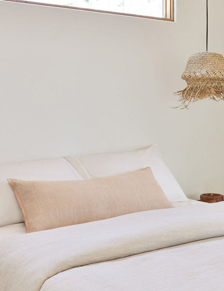 #color::blush #style::extra-long #fill type::with-feather-down-insert #fill type::pillow-cover-only | The arlo Blush pink long lumbar pillow lays on a bed with ivory linens with a jute pendant light hanging next to it