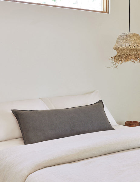 #color::conifer #style::extra-long #fill type::with-feather-down-insert #fill type::pillow-cover-only | The arlo Conifer gray long lumbar pillow lays on a bed with Ivory linens with a jute pendant light hanging next to it
