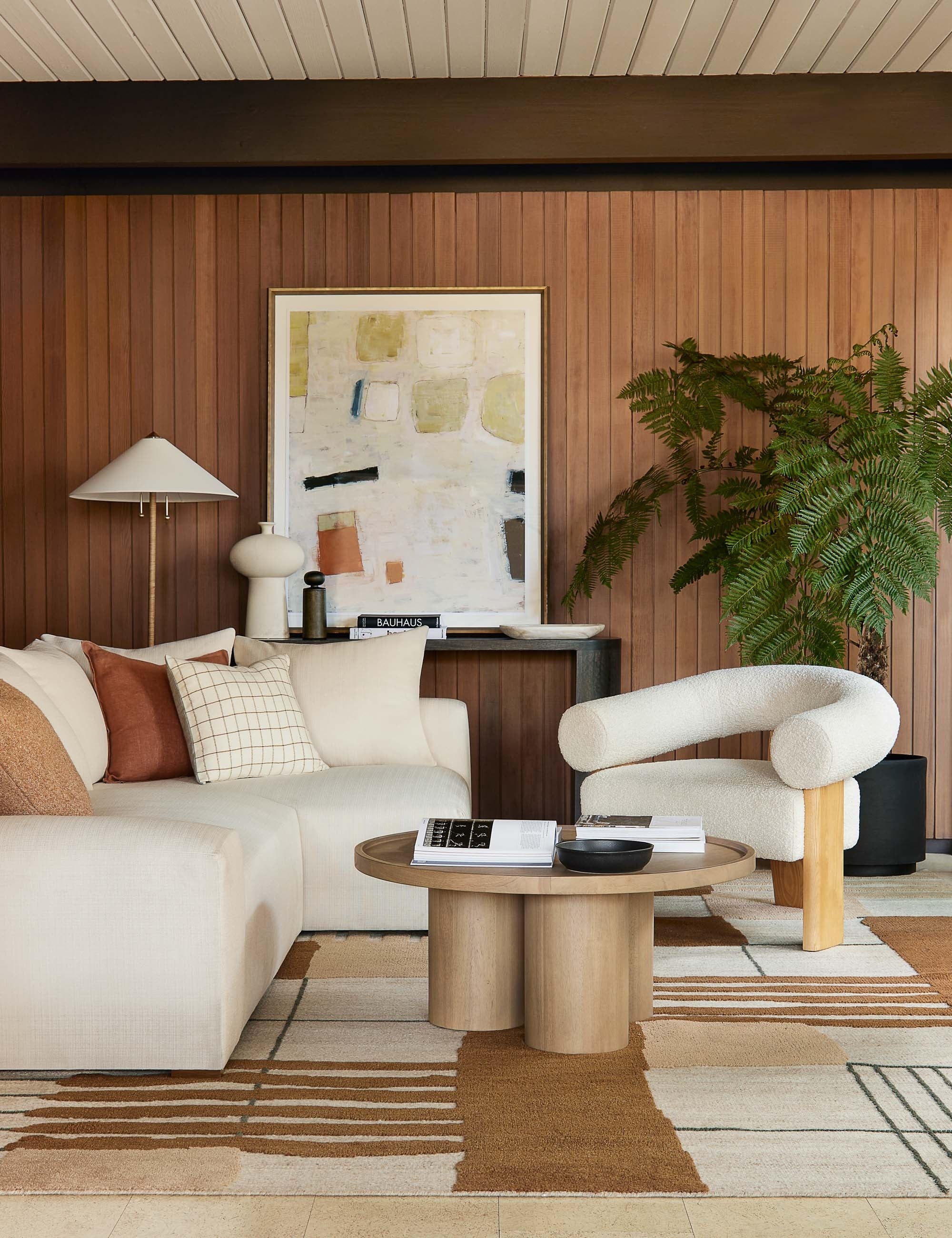The Zora floor lamp with polished base and brass knob detailing sits in a retro living room with wood paneled walls, a sculptural boucle chair, and a circular wooden coffee table