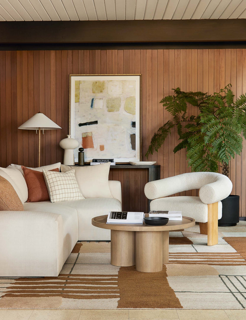 | The Zora floor lamp with polished base and brass knob detailing sits in a retro living room with wood paneled walls, a sculptural boucle chair, and a circular wooden coffee table
