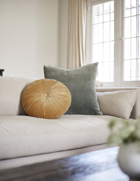 #color::mustard | The Monroe mustard yellow velvet round pillow sits on a natural sofa with a shale blue velvet throw pillow