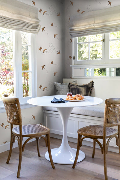 #color::rust | The Sparrow brown wallpaper by Rylee and Cru is in a dining nook with a white sculptural dining table, brown woven chairs that are surrounded by large colonial style windows.