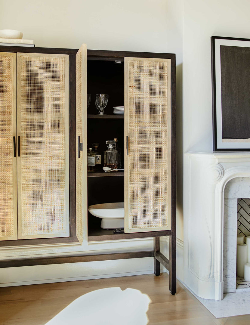 #color::black-and-natural | The Hannah black mango wood cabinet with cane doors sits in a living room with one door open holding a white centerpiece bowl and hosting serveware.