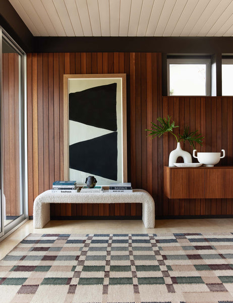 #showall::43011499786339 | The Tate cream boucle upholstered bench sits in a retro entry way with a patterned rug in front of a wood paneled wall and black and white wall art.