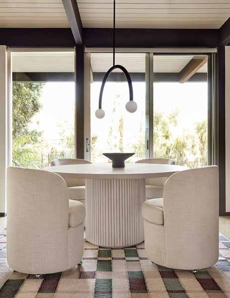 #color::white-wash | The Rutherford white-washed acacia wood round dining table with pedestal base sits in a dining room surrounded by four linen dining chairs atop a beige patterned carpet with red and green accents.