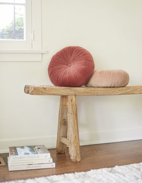 #color::coral | The Monroe coral pink velvet round pillow sits on a wooden bench with the rosewater pink round velvet pillow
