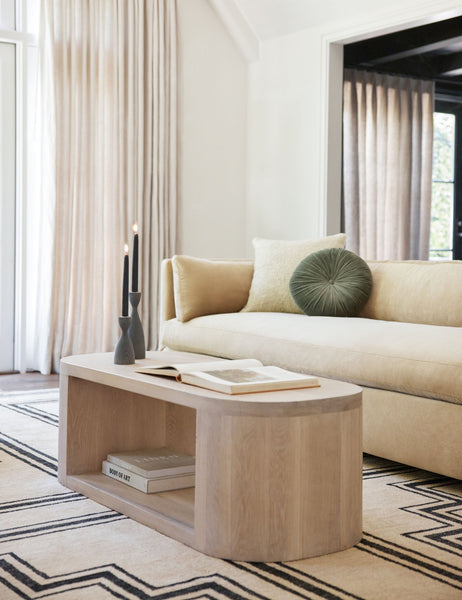 #color::moss | The Monroe moss green velvet round pillow sits on a natural sofa in a living room with an oval coffee table and geometric rug