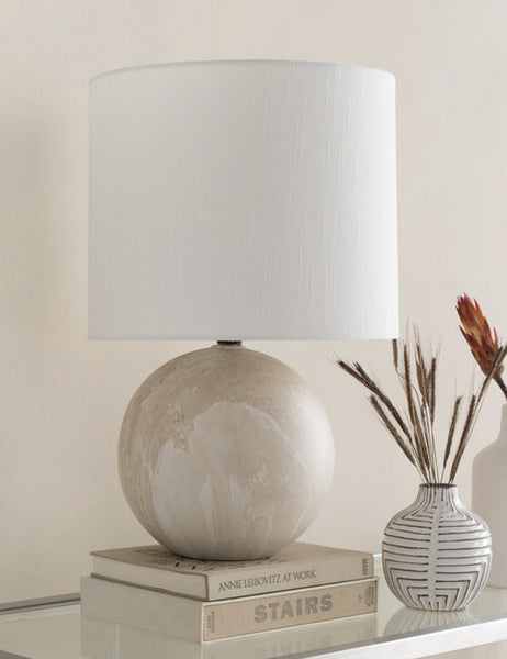 #color::tan | The Vivienne tan ceramic table lamp with spherical base sits atop a glass sideboard with a white vase and a stack of books